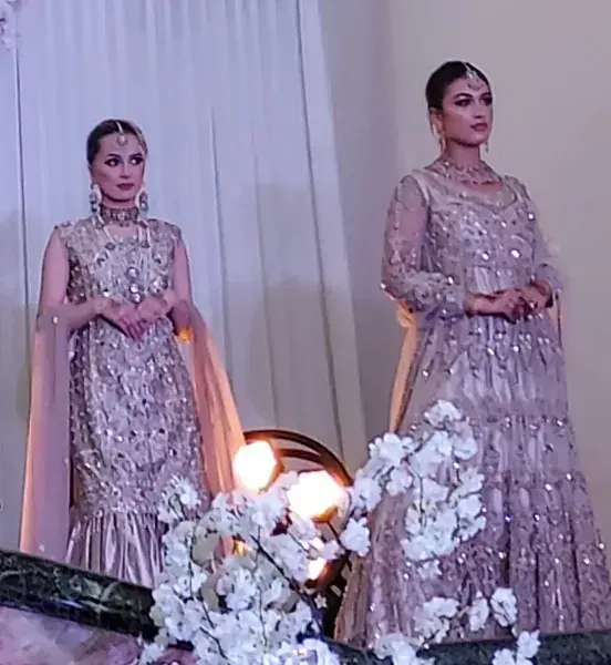 Two women in shimmering lavender gowns stand on a runway beside white floral arrangements.