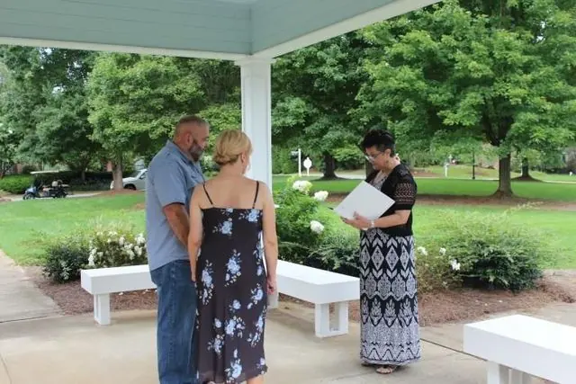 Three people stand under a pavilion; one reads from a paper in a park.