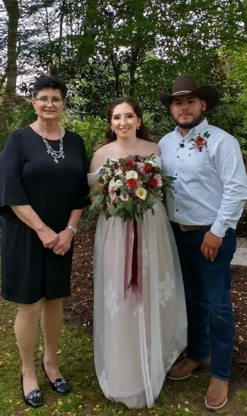 Bride in white dress holding bouquet stands with two guests outdoors in front of trees.