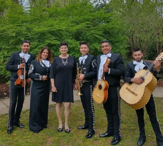 Six musicians in black formalwear pose outdoors with violins and guitars in a green park.