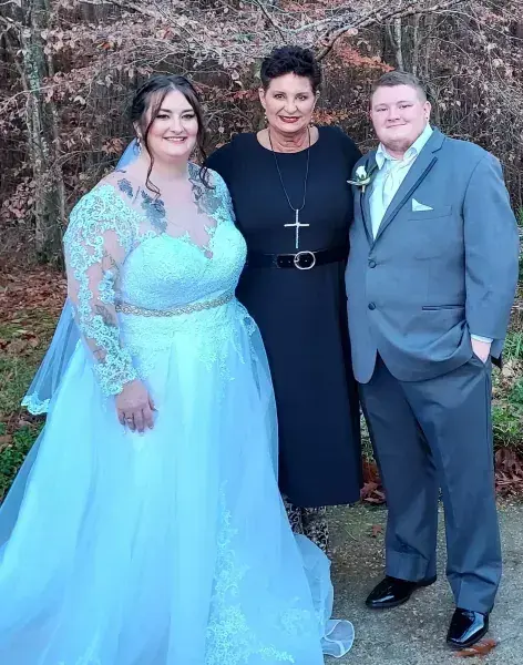 Three people posing outdoors; one in a light blue gown, one in black with a cross, one in a gray suit.