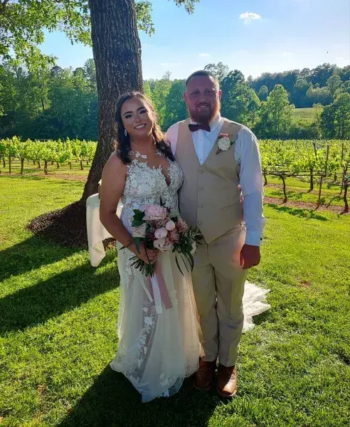 Wedding couple posing outdoors in sunlight, bride holding bouquet beside groom in a tan suit