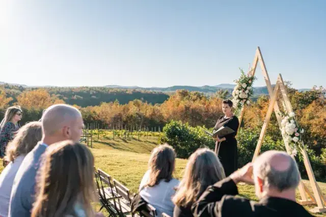 Outdoor wedding ceremony with seated guests, floral arch, and speaker in front of autumn vineyard scenery