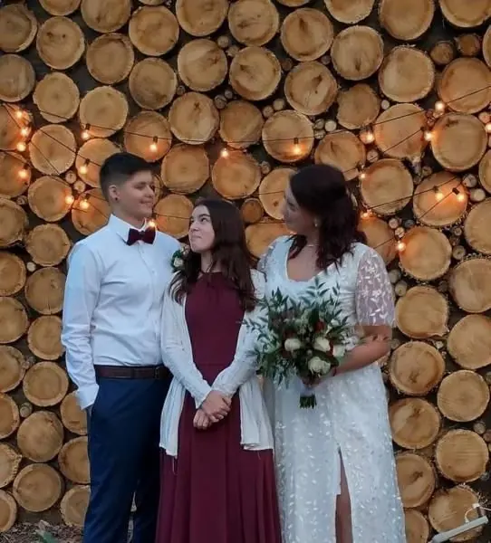 Three people in formal attire posing in front of a wall of stacked logs with string lights