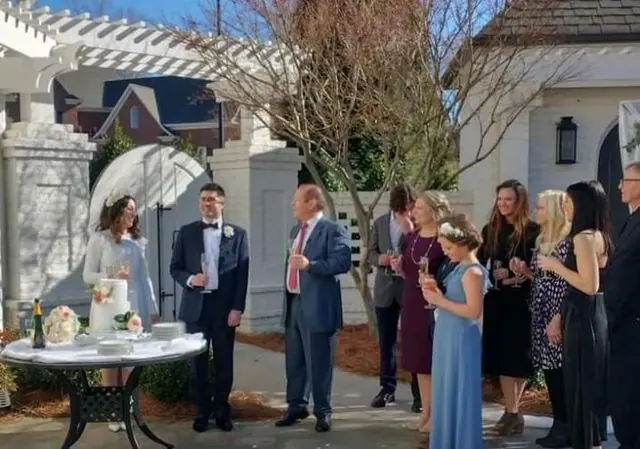 People mingling at an outdoor gathering by a white house and pergola, holding drinks and chatting.