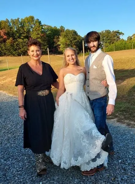 Three people posing outdoors in formal attire, with a bride in a white wedding dress centered between two companions.