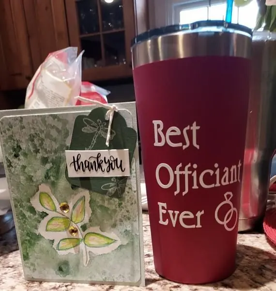 Thank-you card beside red tumbler reading “Best Officiant Ever” on a kitchen counter