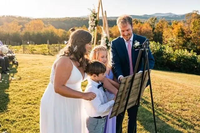 Wedding party reading from a music stand outdoors at sunset in a grassy field, with mountains in the background