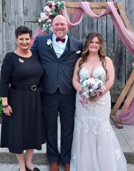 Three people posing at a wedding, with the bride holding a bouquet in a white gown and pink drapes behind them.