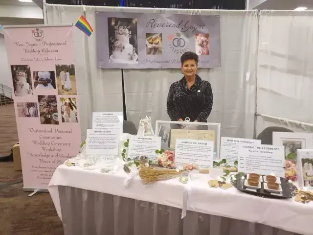 Woman at a memorial display booth with photos, flowers, candles, and handwritten signs behind a white table.