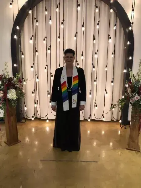 Person standing in a black gown with a rainbow stole before a lit draped backdrop and floral arrangements