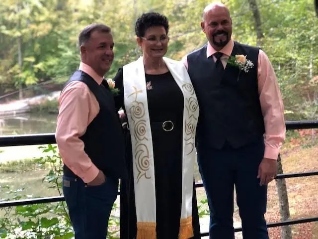 Three people posing together outdoors by a railing in front of a wooded stream.