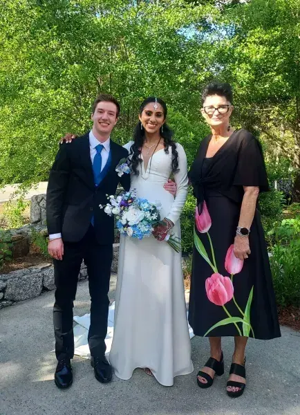 Three people pose outdoors in formal attire, with a bride holding a blue and white bouquet.