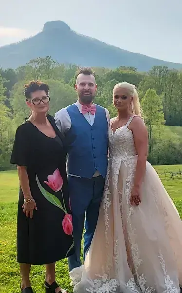 Three people pose in wedding attire outdoors with a mountain backdrop.
