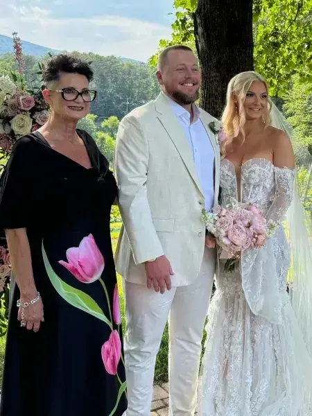Three people posing outdoors at a wedding, with the bride in a white lace gown holding pink flowers.